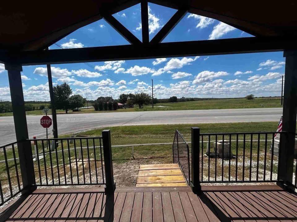 View from a shaded porch with wooden steps, overlooking a quiet rural road, open fields, and a bright blue sky dotted with fluffy white clouds. Calm atmosphere.