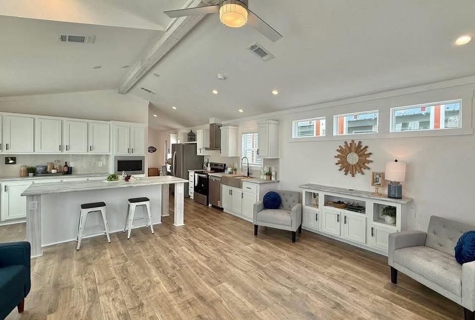 Spacious, modern kitchen and living area with white cabinets, wood flooring, and gray seating. Bright with natural light from large windows.