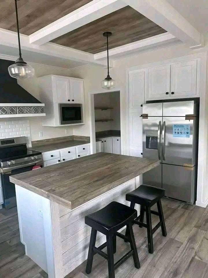 Modern kitchen with white cabinets, stainless steel appliances, wood countertops, and two black stools at an island. Pendant lights hang from the ceiling.