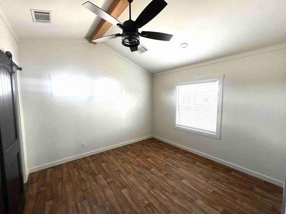 Empty room with white walls and wood floor. Ceiling features a black fan, exposed beam, and recessed lighting. Light enters through two windows.