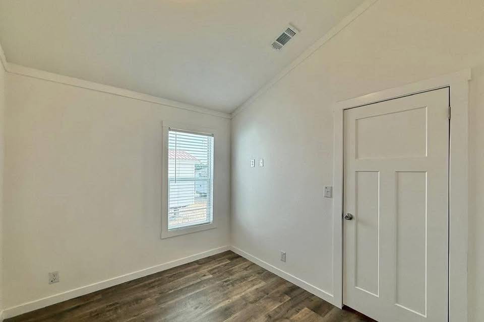 Small, empty room with light-colored walls and wooden floor. A single window with blinds on the left, and a closed white door on the right. Calm and simple atmosphere.