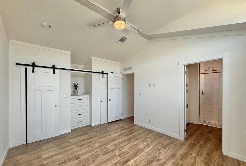 A minimalist white room with light wood flooring, featuring a ceiling fan. A closet with sliding barn doors is on the left, and an open doorway leads to a bathroom on the right.
