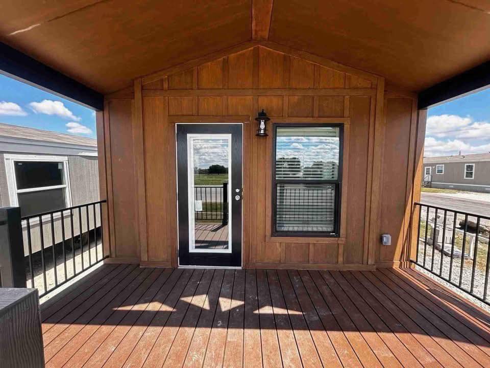 Wooden deck area with dark railing, featuring a central glass door and window framed in black. A vintage lantern is beside the door. Sunny skies above.
