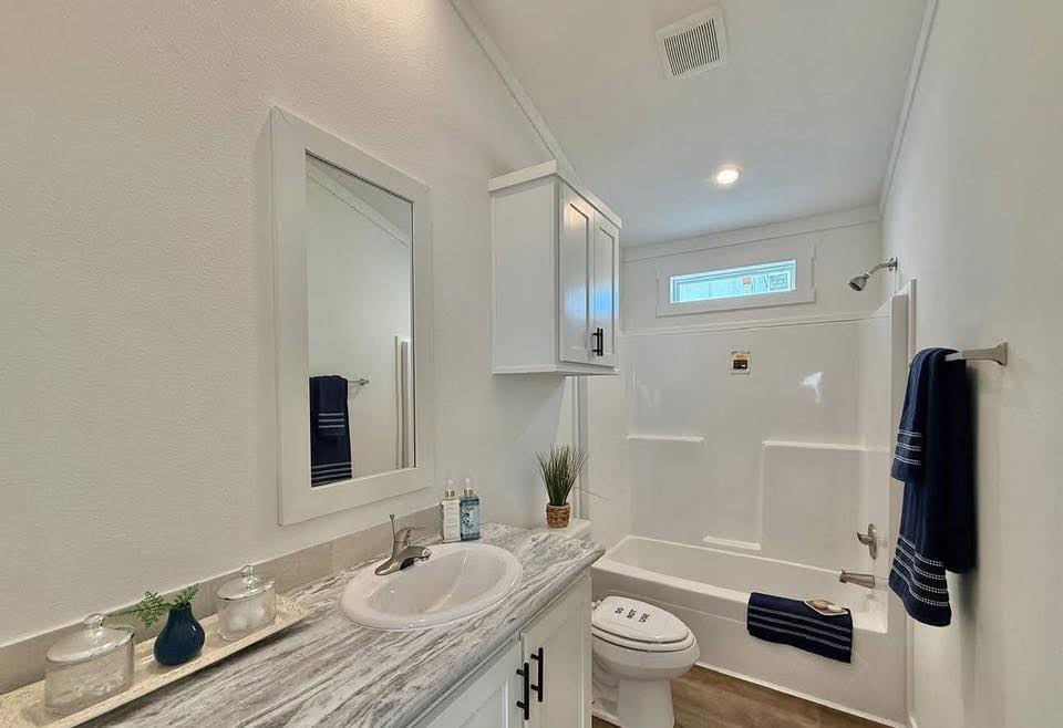 Bright bathroom with a white vanity and marble countertop. Wall-mounted mirror above the sink, blue towels, decorative plants, and a bathtub with a shower. Clean and modern feel.