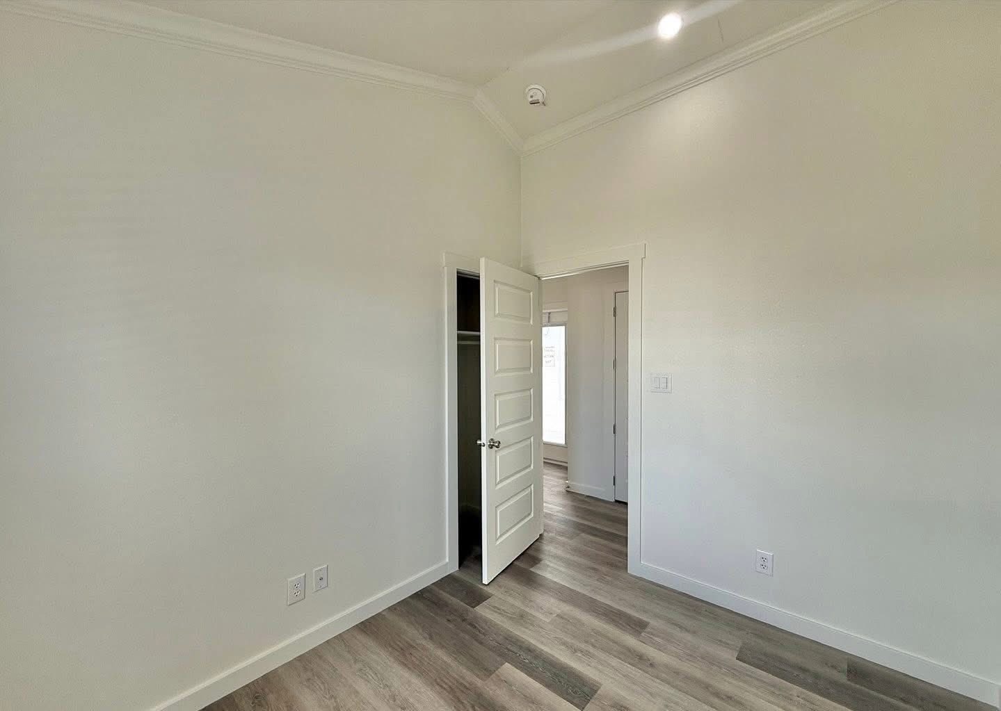 Minimalist empty room with light-colored walls and wood flooring. An open door reveals a small closet, and another doorway leads to a well-lit hallway.