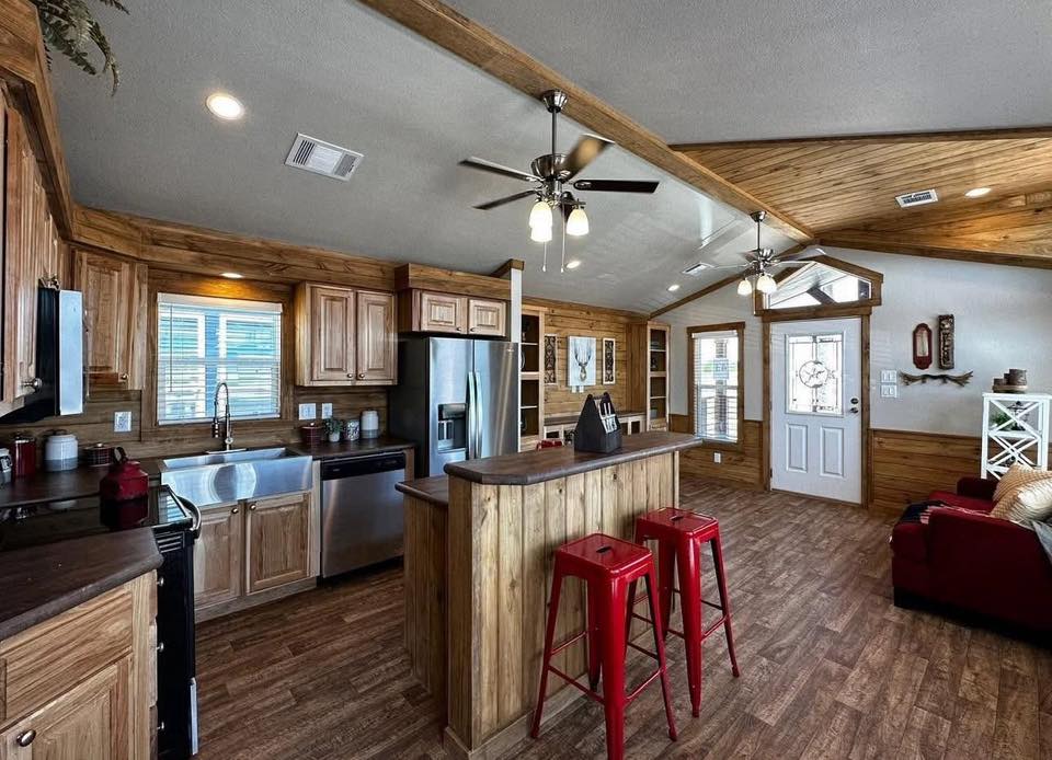 Cozy rustic kitchen and living area with wooden cabinetry, stainless steel appliances, and a small island with two red stools. Ceiling fans and red sofa add warmth.