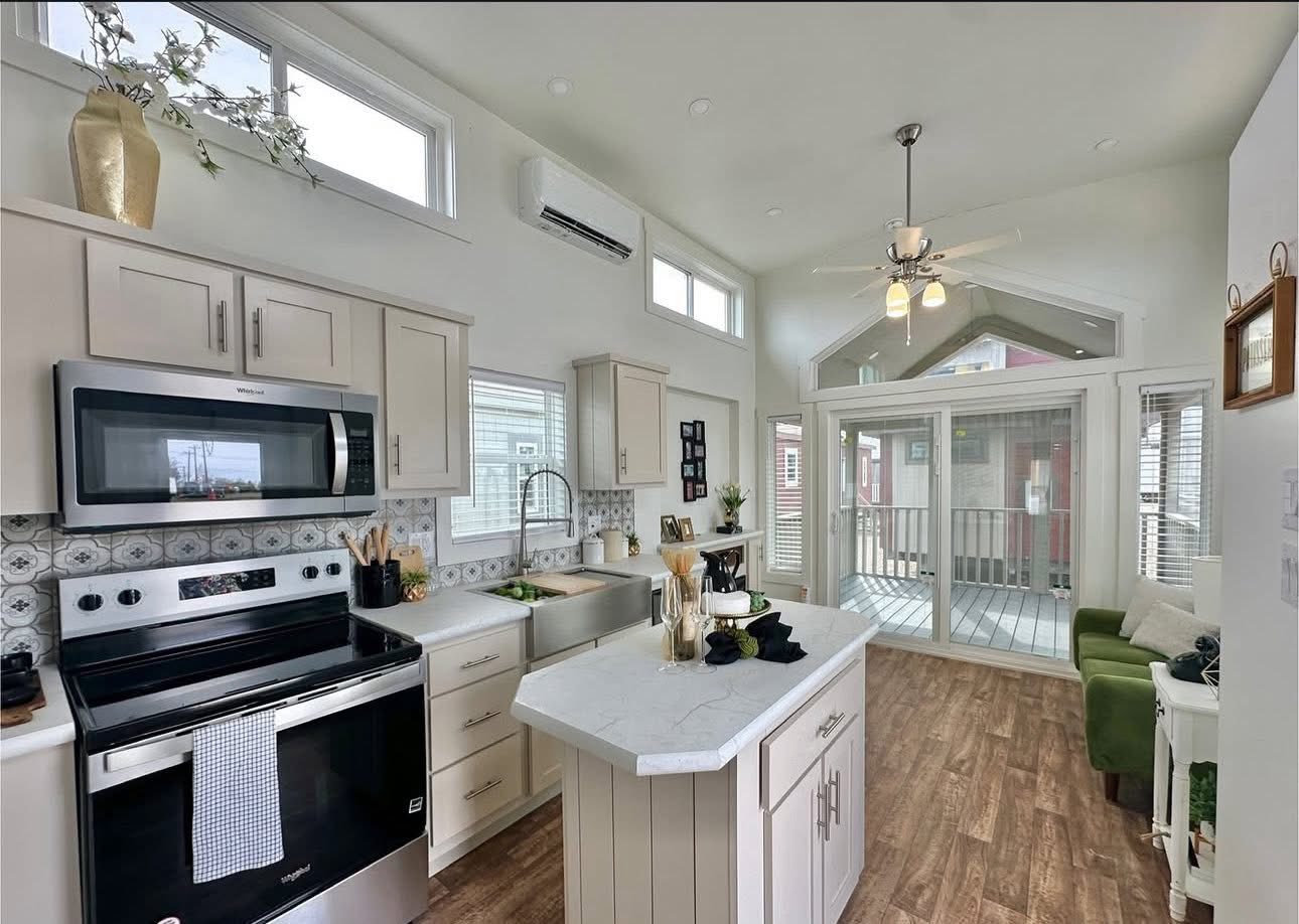 Bright, airy kitchen with modern appliances, white cabinetry, and a central island. The room opens to a sunlit seating area with large windows.