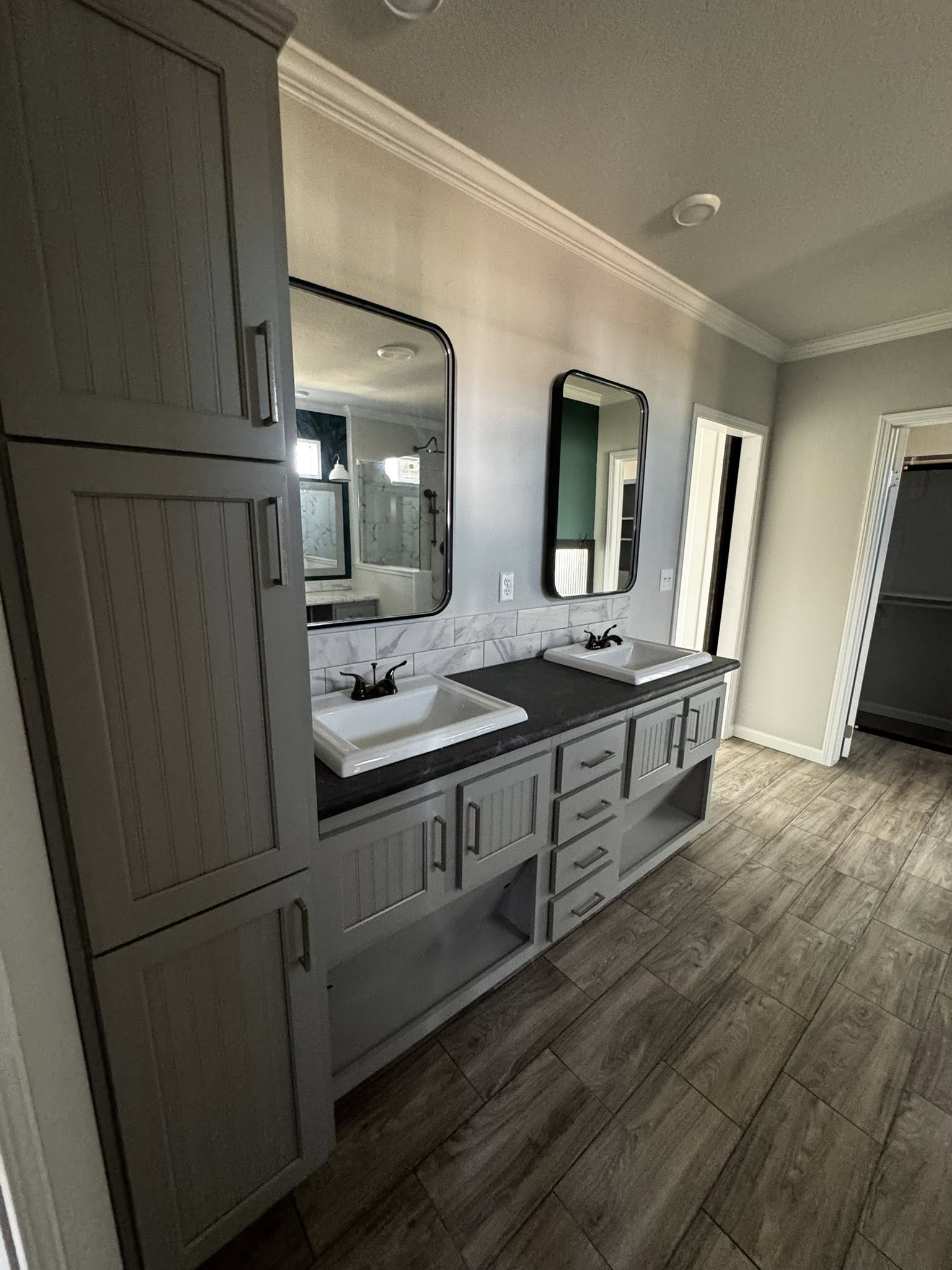 Modern bathroom with dual sinks and black fixtures on a dark countertop. Two mirrors hang above, framed by light gray cabinetry. Wood-patterned tile flooring creates a warm ambiance.