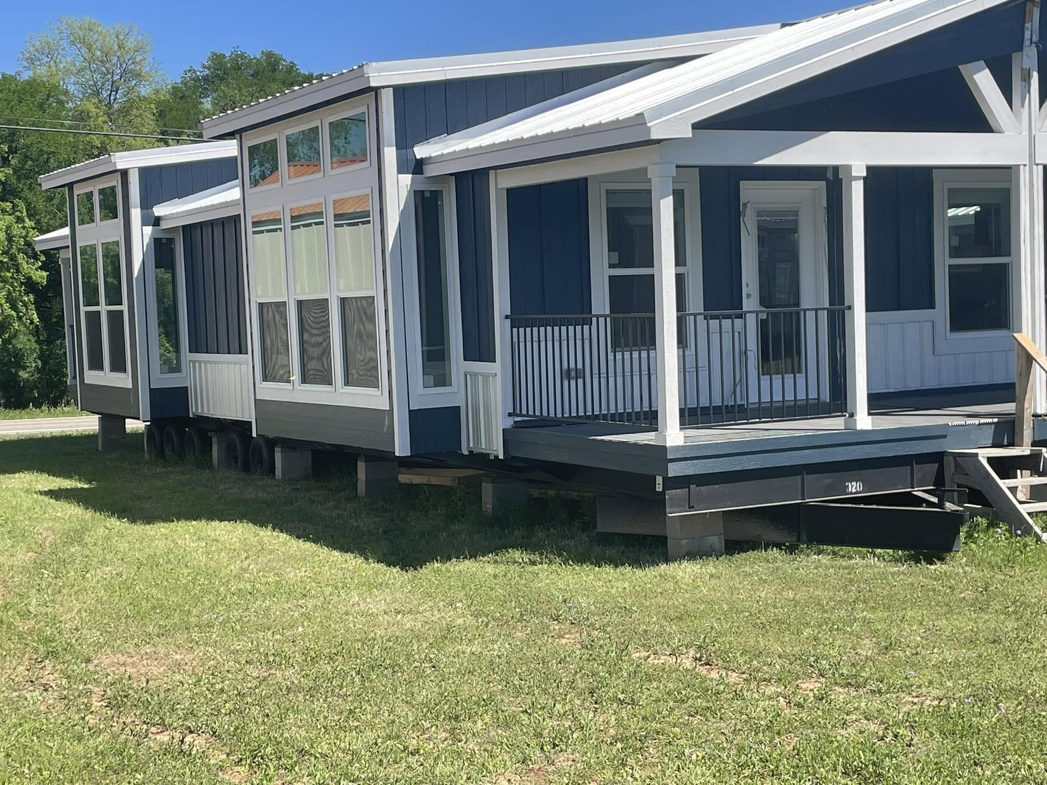 A blue and white modular home with large windows and a small porch is set on a grassy lawn. The sunny day highlights the modern design.