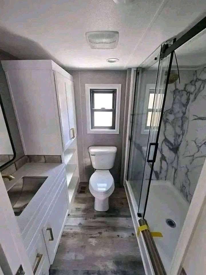 Small, modern bathroom with white cabinets, a toilet, and a glass-enclosed shower with marble tiles. Natural light from a window brightens the room.