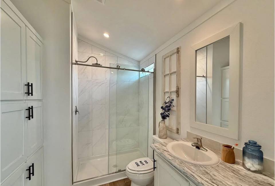 Small, bright bathroom with a marble sink, glass shower, and white cabinetry. There's a sign on the toilet reading "Do Not Use" and a vase with purple flowers.