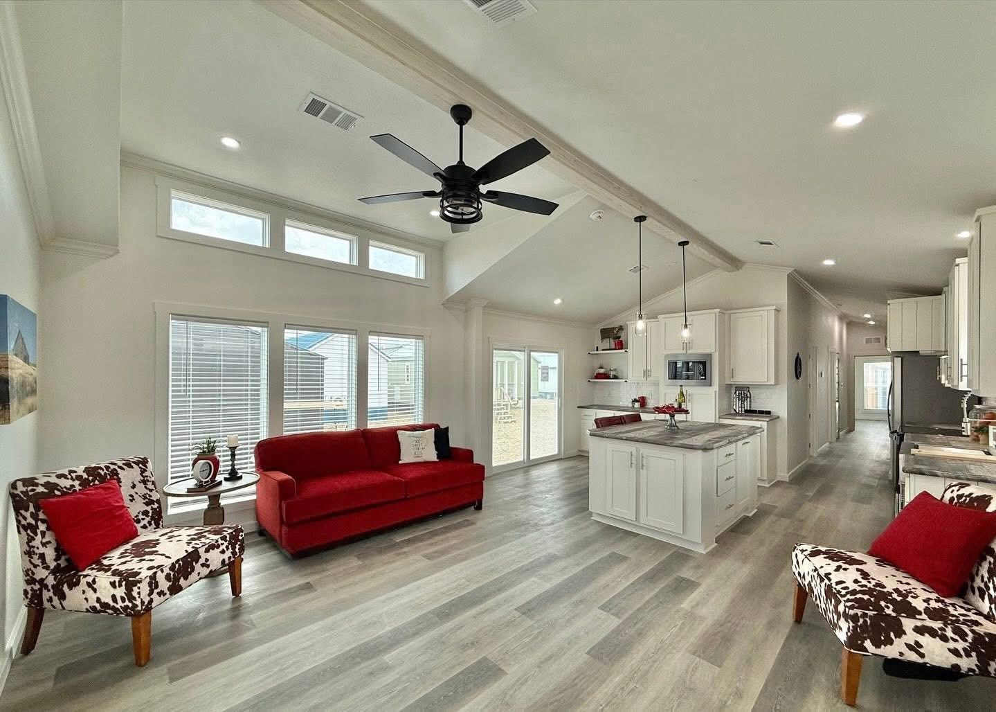 Spacious, modern living room and kitchen. Features red sofa, cow-print chairs, vaulted ceiling, ceiling fan, white cabinets, and warm wood flooring. Cozy vibe.