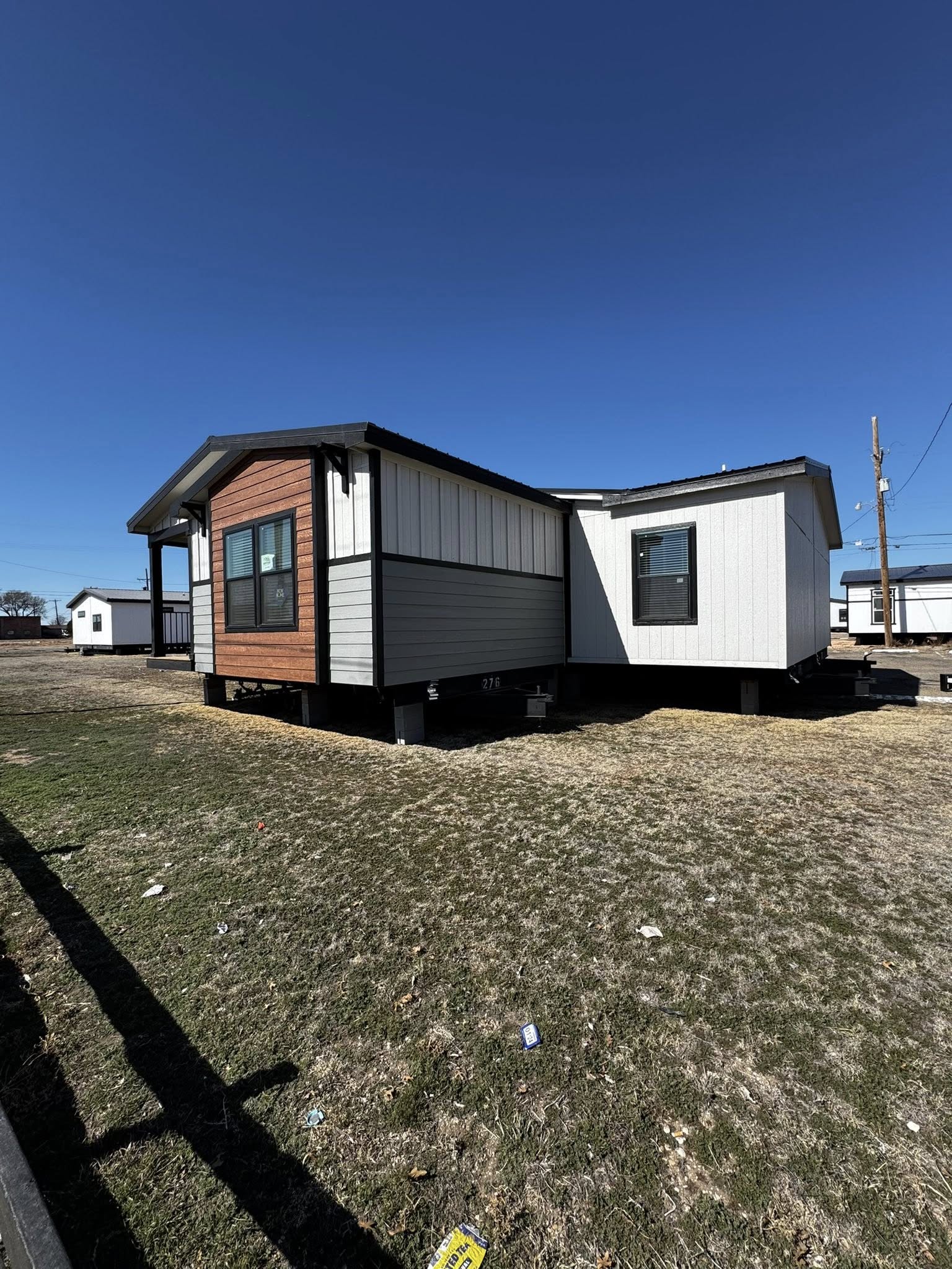 Modern mobile home with brown and gray siding on a grassy lot under a clear blue sky. It stands elevated on a foundation, casting a shadow.