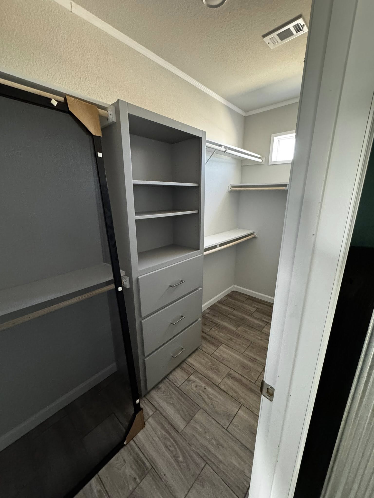 Small walk-in closet with gray shelves and drawers on the left, wooden flooring, and a window to the right. The space is neat and empty.