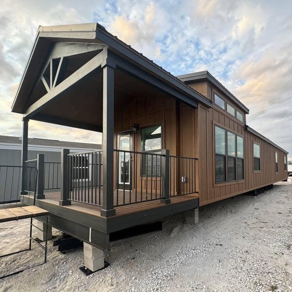 Modern cabin-style mobile home with brown wood siding and a covered porch. The design features large windows and black railings. The sky is partly cloudy.