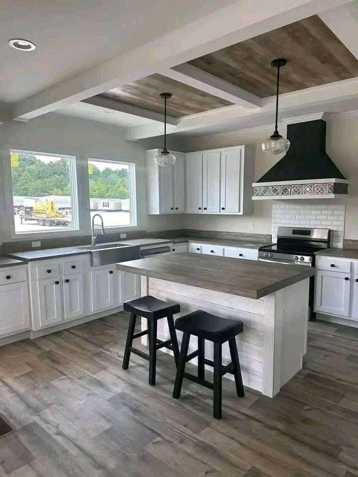 Modern kitchen with white cabinets, a large island with two black stools, and stainless steel appliances. Pendant lights hang from a coffered ceiling.