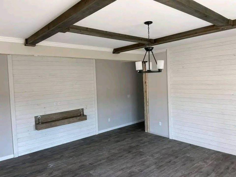 A room with wooden beams on the ceiling, gray walls, and white wood paneling. A dark chandelier hangs from the center, creating a rustic yet modern feel.