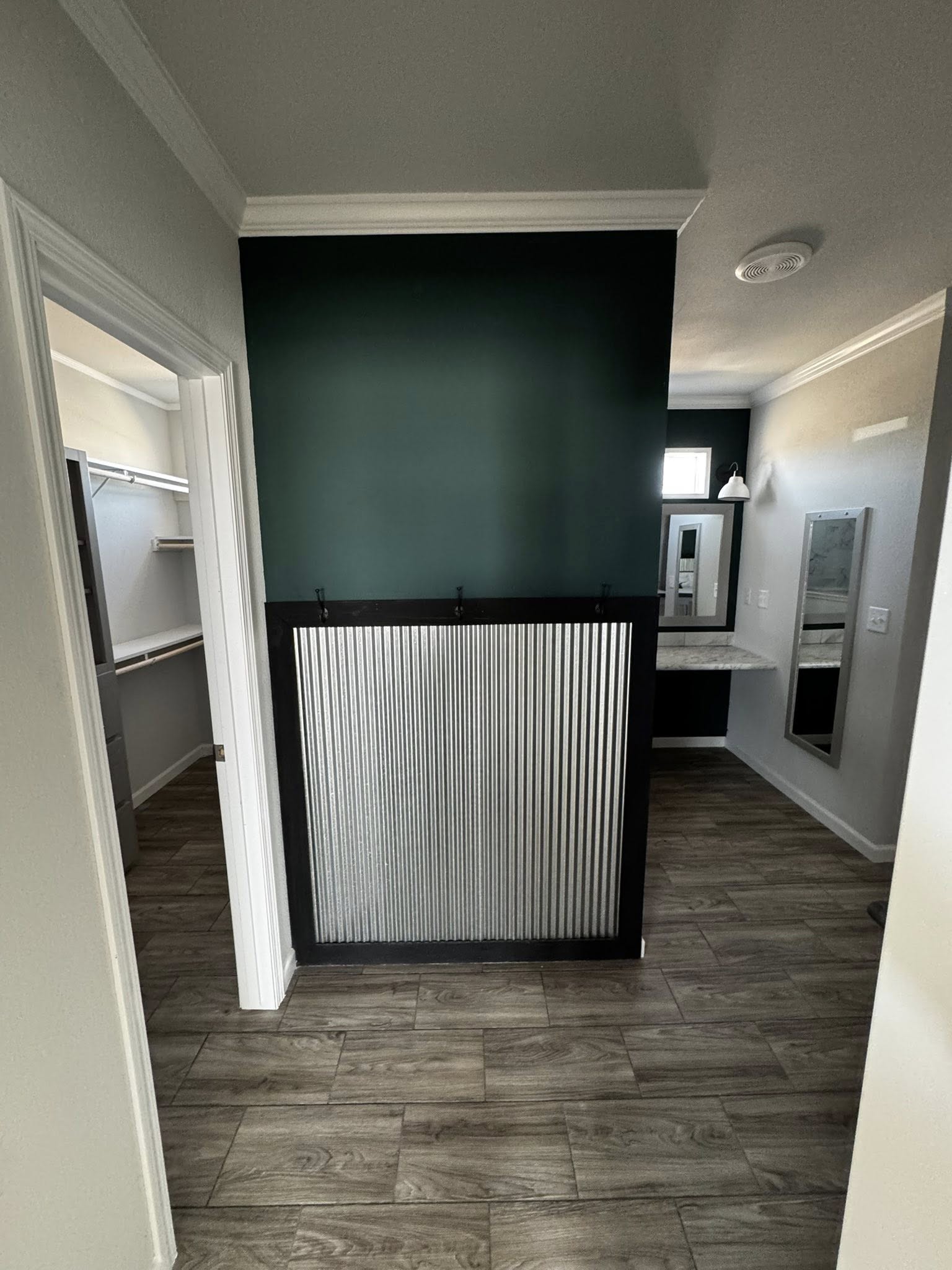 View of a modern room with wood-textured tile flooring, a green accent wall featuring a metal paneling divider, and adjacent open closet and vanity.