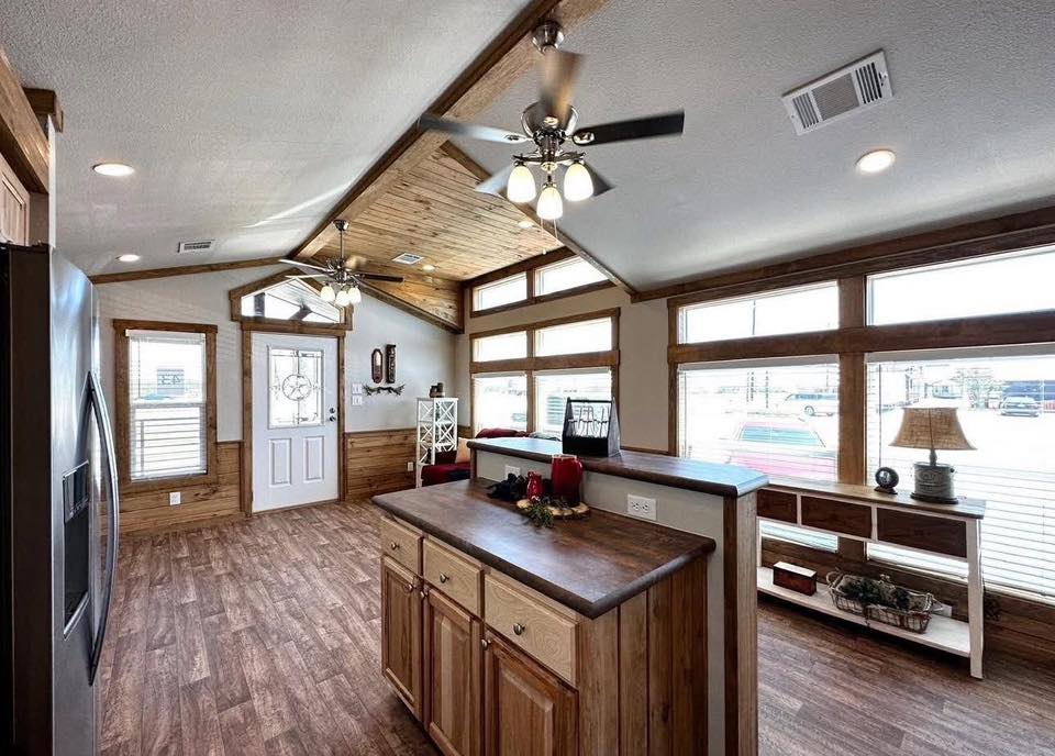Spacious rustic kitchen with wooden floors and cabinets. Natural light streams through large windows. Ceiling fans add a cozy, inviting feel.