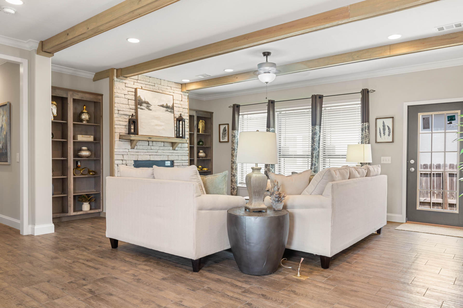 Spacious living room with beige sectional sofa, stone fireplace, and wooden shelves. Large windows with curtains allow natural light. Cozy and inviting atmosphere.