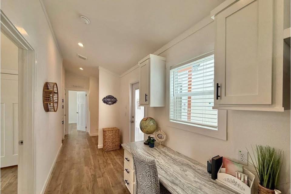 A cozy hallway with wooden flooring and white walls features a window with blinds, a desk with a globe, cabinets, and a wicker basket, creating a warm, inviting ambiance.
