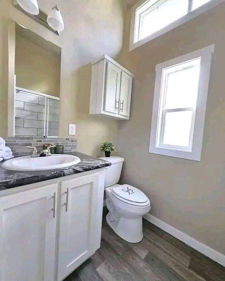 Compact bathroom with beige walls and wood flooring. Features a vanity with a black countertop, white cabinets, toilet, and small window, creating a clean, bright ambiance.