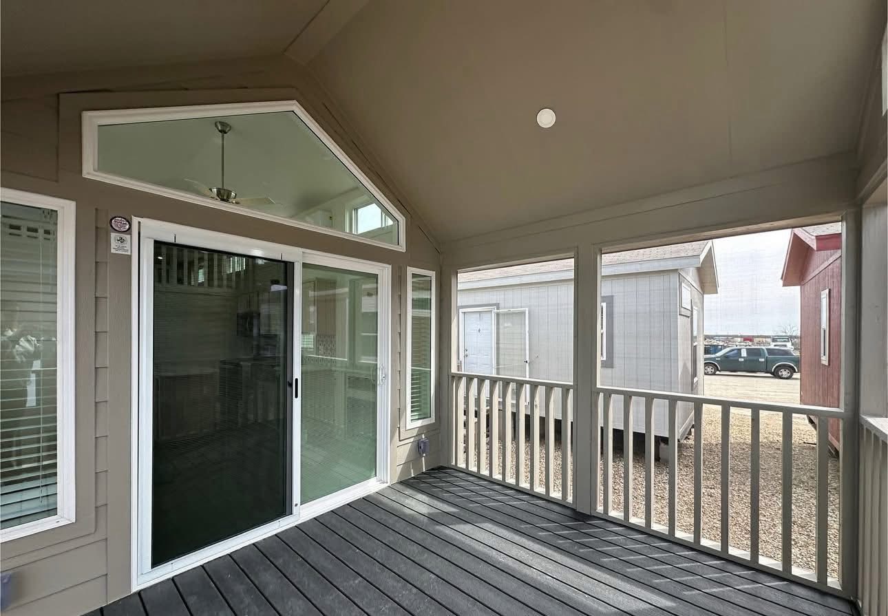 Covered porch with white railings and a sloped ceiling, featuring large sliding glass doors and triangular windows. Nearby structures and cars are visible outside.