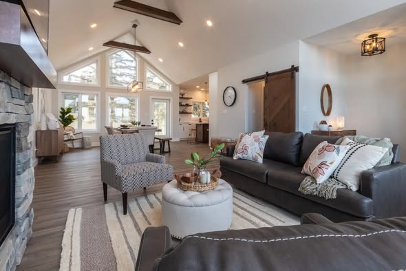 Spacious, cozy living room with vaulted ceiling, large windows, a stone fireplace, gray sofa, patterned chair, and stylish decor. Bright, inviting ambiance.