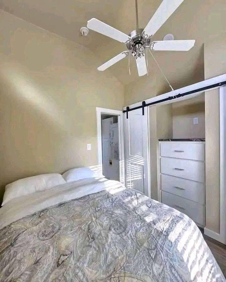 Compact bedroom with a bed covered in a patterned duvet, a white ceiling fan above, and a sliding door revealing a closet with drawers. Bright, airy atmosphere.