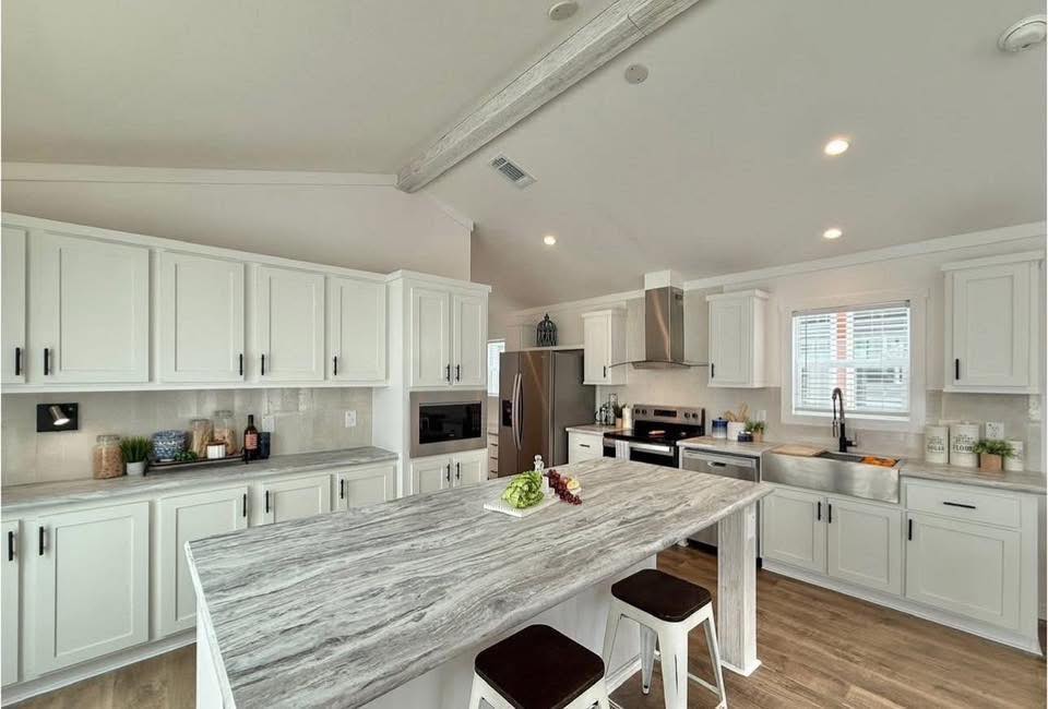 Modern kitchen with white cabinets, stainless steel appliances, and a large gray marble island. Bright lighting, wooden floors, and a cozy atmosphere.