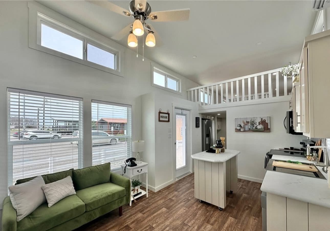 Modern kitchen with light wood flooring and white cabinets. Features a central island, stainless steel appliances, and a cozy fireplace. Bright, airy loft above with framed photos and decorative accents, exuding a warm, inviting atmosphere.