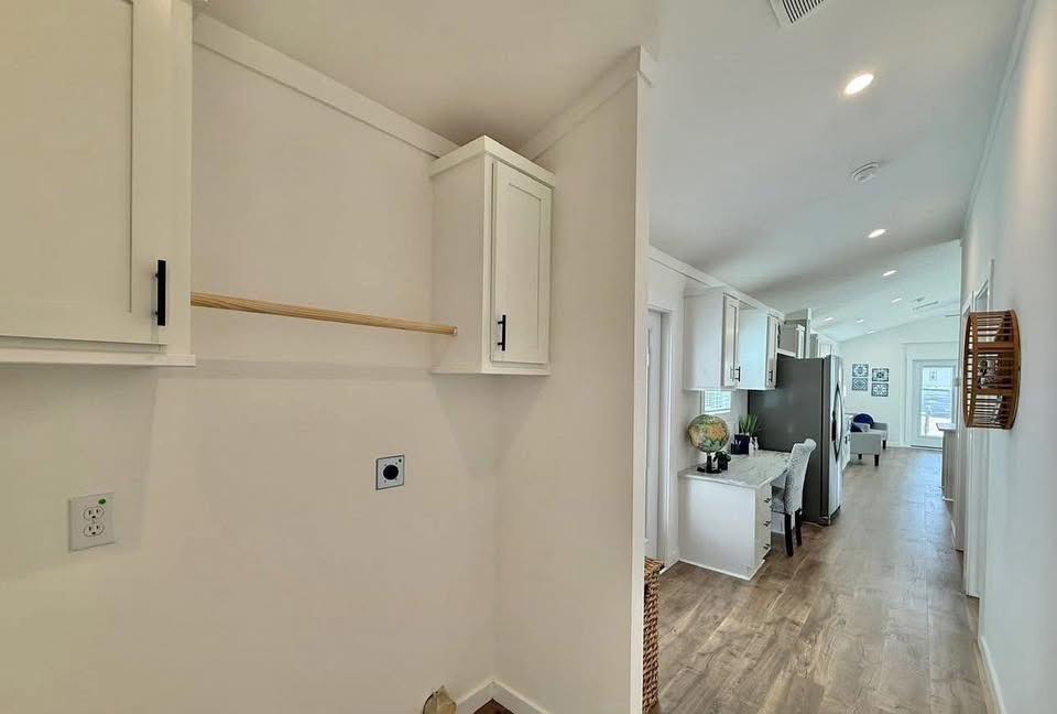 Narrow hallway with light wood floors, white walls, and cabinets. A distant kitchen area with modern appliances and decor is visible, conveying a clean, airy atmosphere.