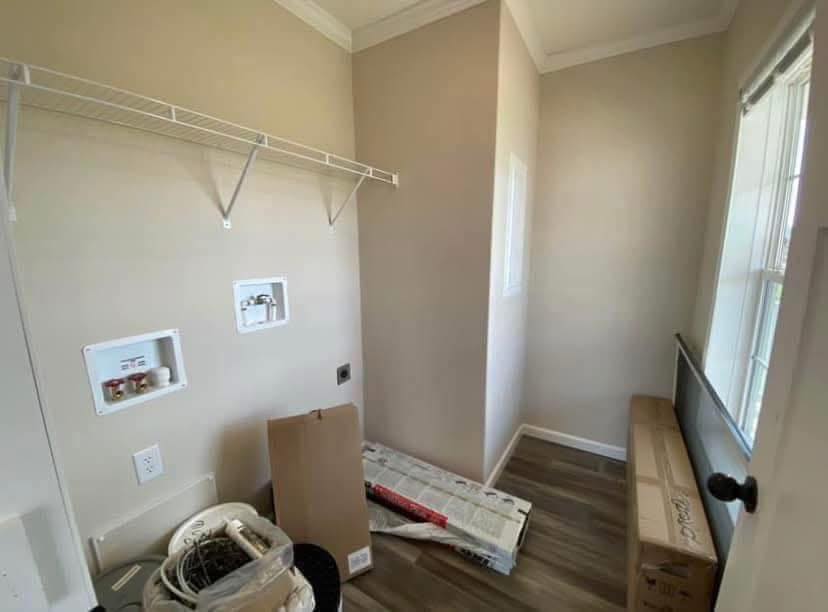 Narrow laundry room with light walls, dark wood floor, empty shelf, and washer hookups. Boxes and packaging on the floor suggest ongoing setup.