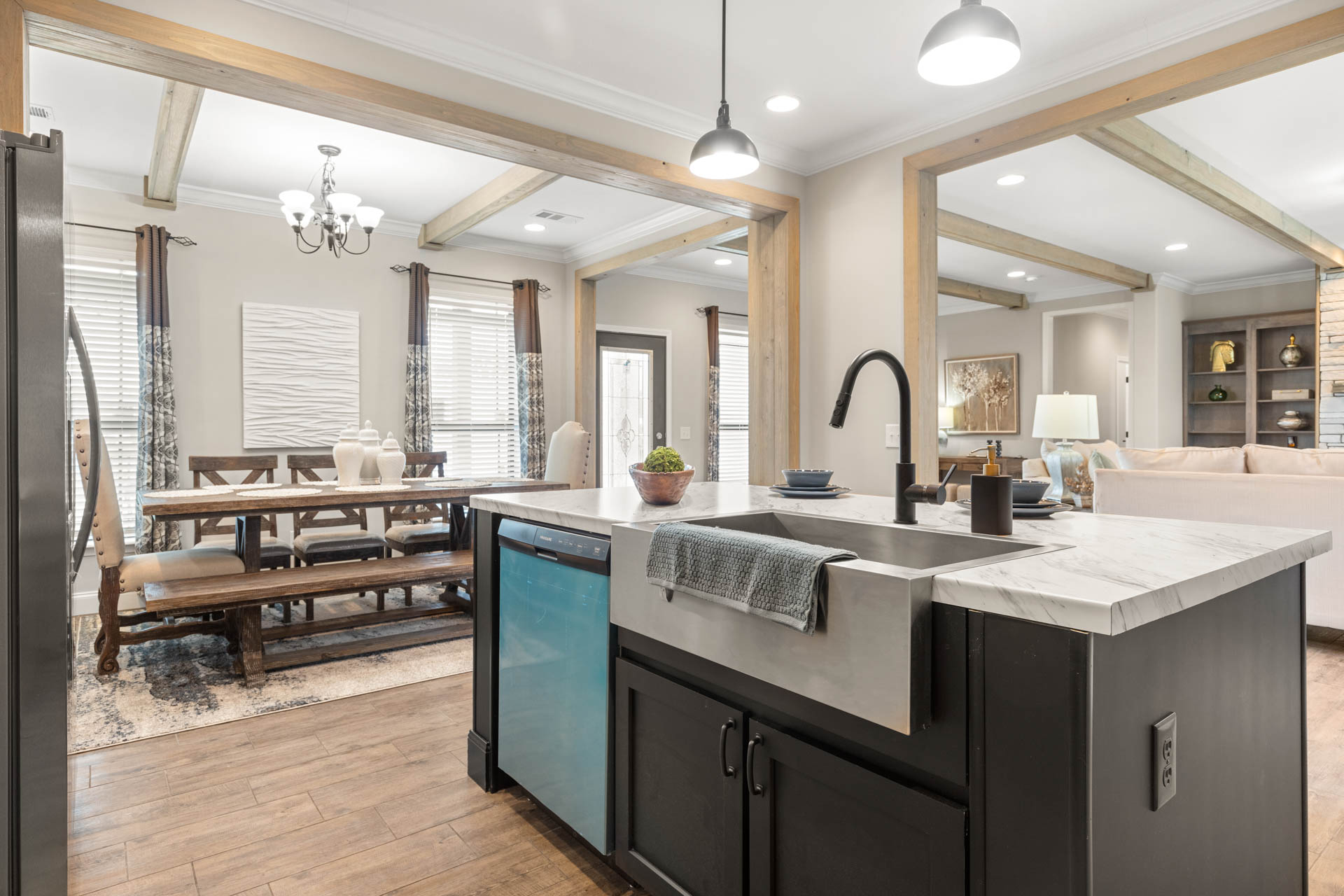 Modern kitchen and dining area with wood beams, light fixtures, and ample natural light. Features marble island, farmhouse sink, and wooden dining table. Cozy and elegant ambiance.