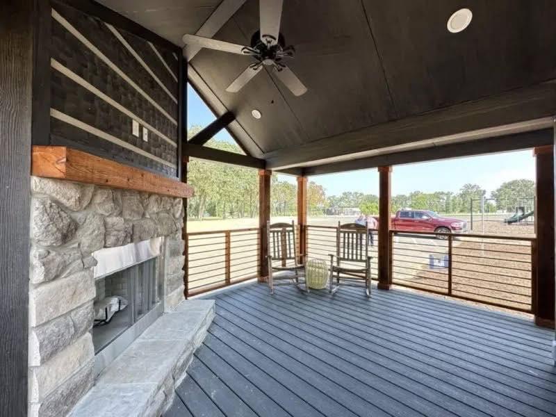 Covered patio with a stone fireplace, two rocking chairs, and ceiling fan. Overlooks a grassy field with trees and a red truck. Cozy, rustic setting.