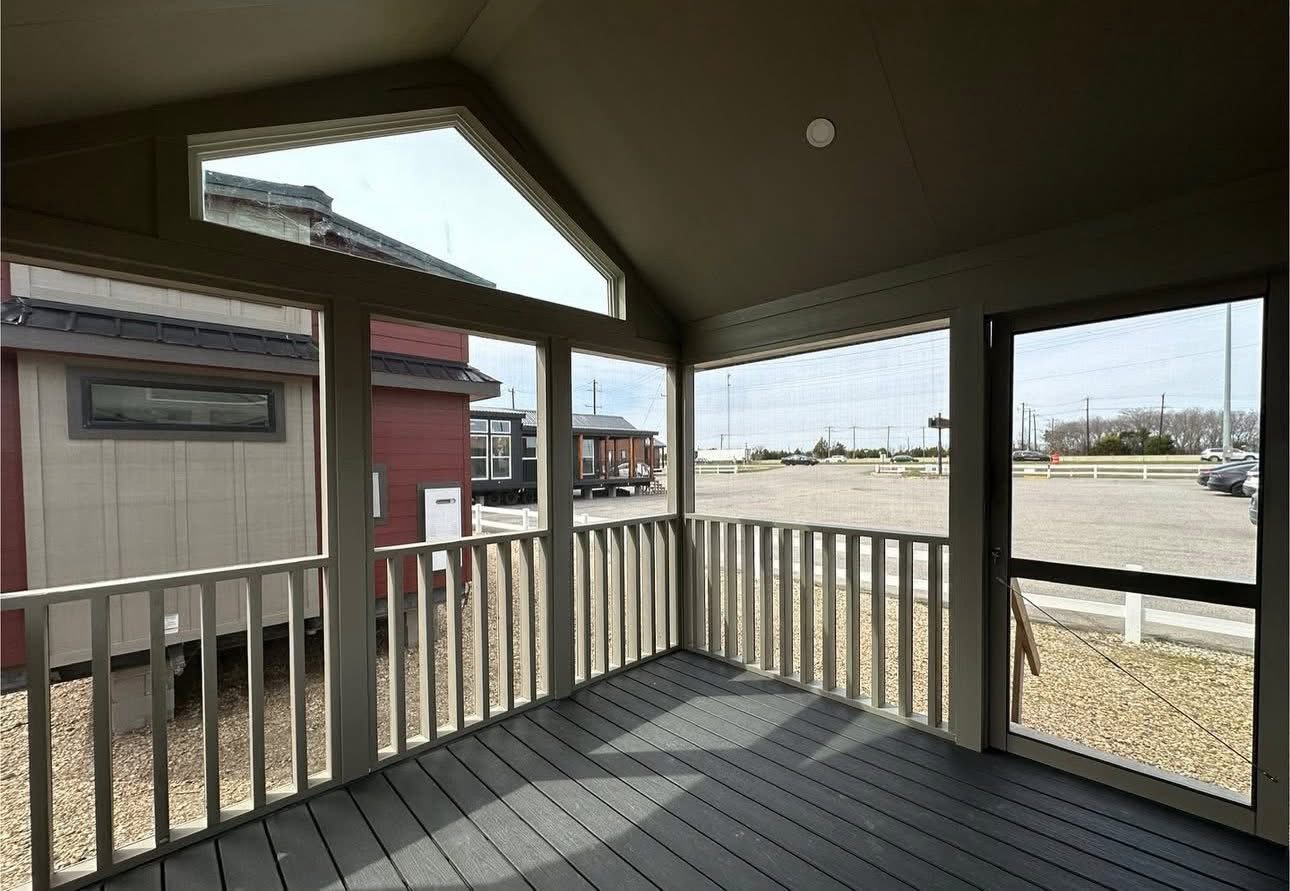 View from a screened porch with white railings overlooking a gravel lot. Tiny homes are visible outside, under a bright sky, creating a serene setting.