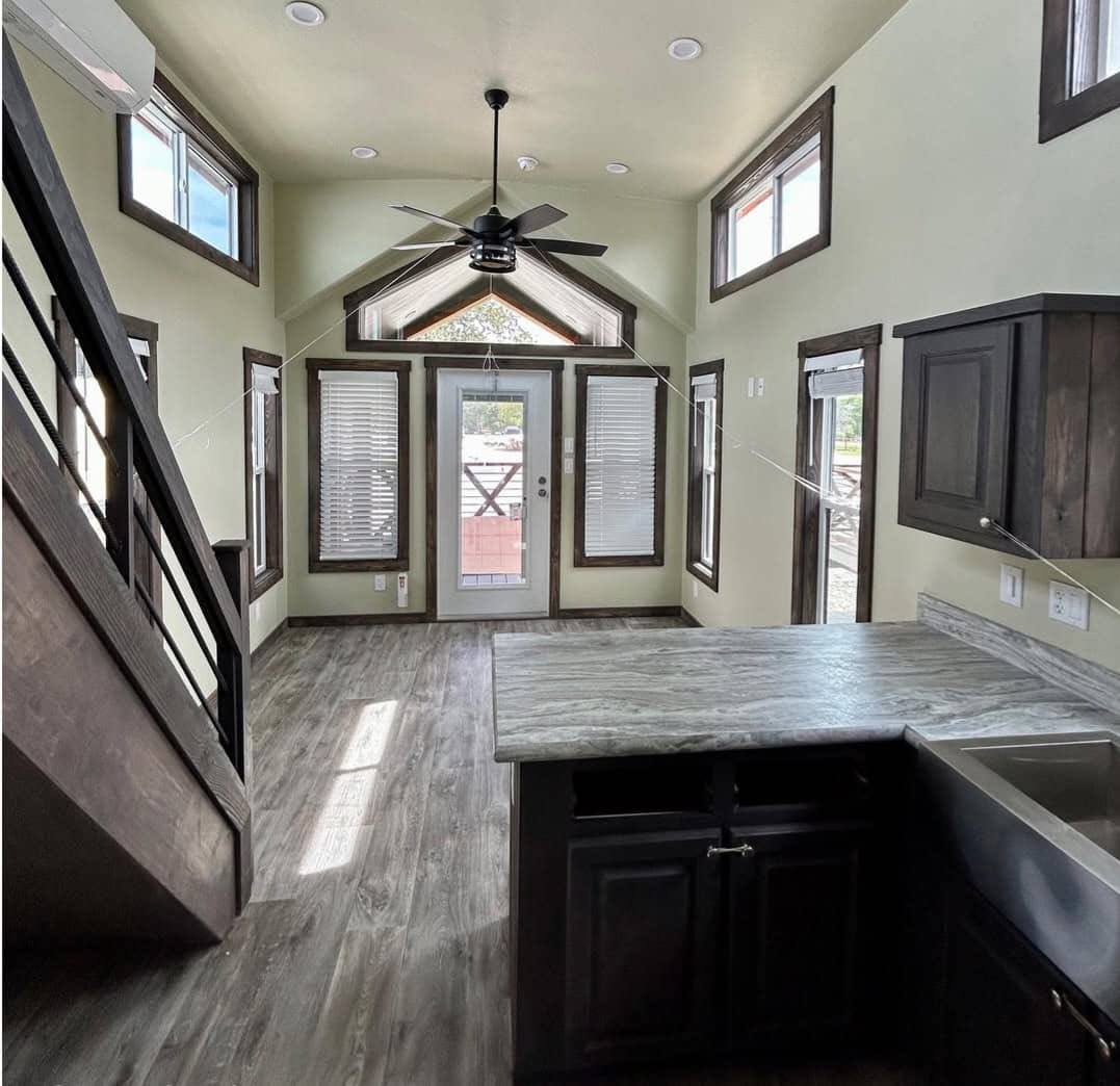 Interior of a modern tiny home with high ceilings, pale walls, dark wood accents, and a loft. Natural light streams through large windows and a glass door.