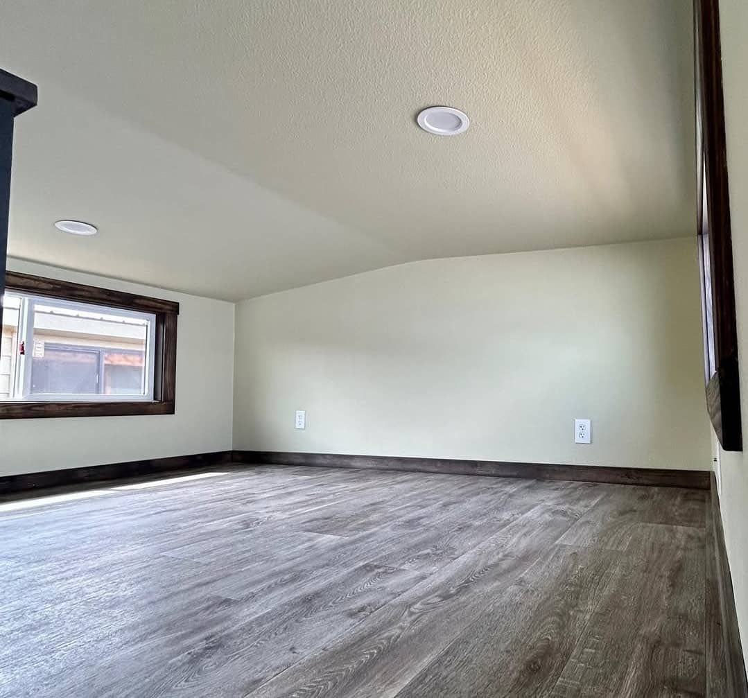 A small, cozy loft space with light wood flooring and softly curved white walls. A single window with dark trim allows natural light to enter.