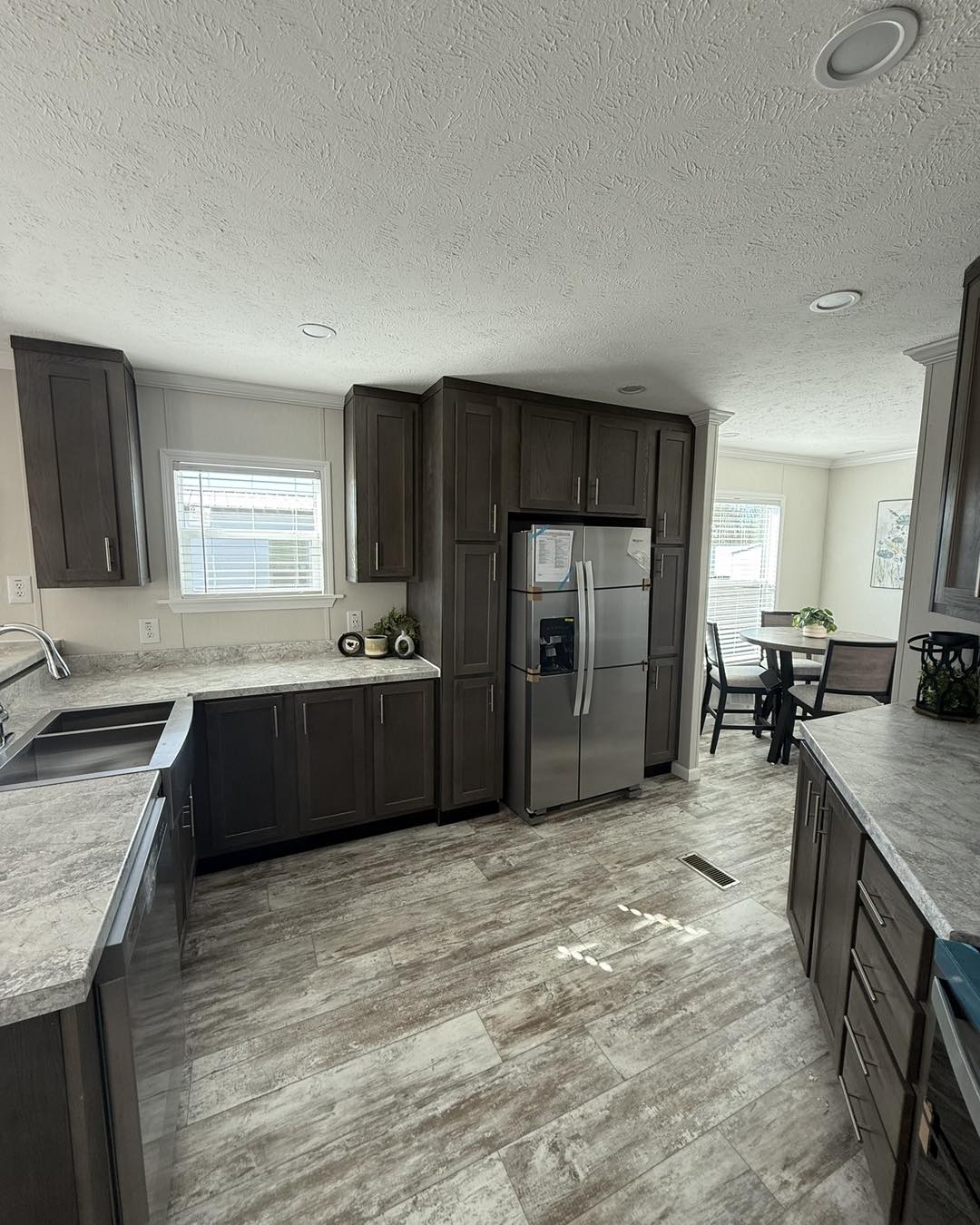 Modern kitchen with dark wood cabinets, stainless steel appliances, and granite countertops. Bright natural light from windows, dining area visible.