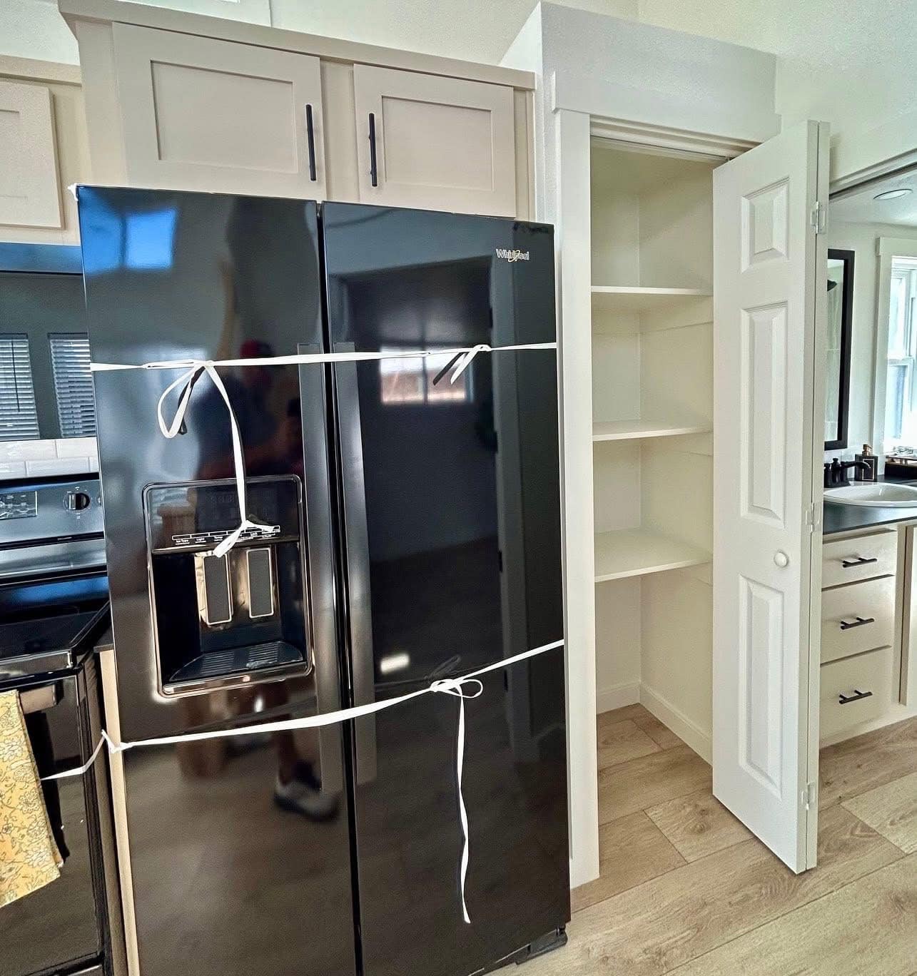 A black refrigerator secured with string stands beside an empty pantry with open white shelves. The kitchen has light wood floors and neutral tones.