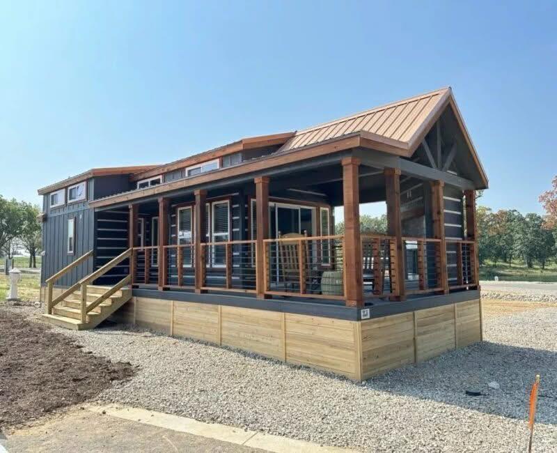 A modern tiny house with a gabled roof, wooden deck, and railing sits on gravel. The exterior is dark with warm wood accents, surrounded by trees under a clear sky.