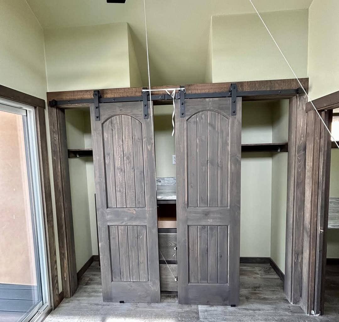 Two rustic sliding barn doors with dark wood finish cover an open closet. The room has pale green walls, a wooden floor, and ample natural light.