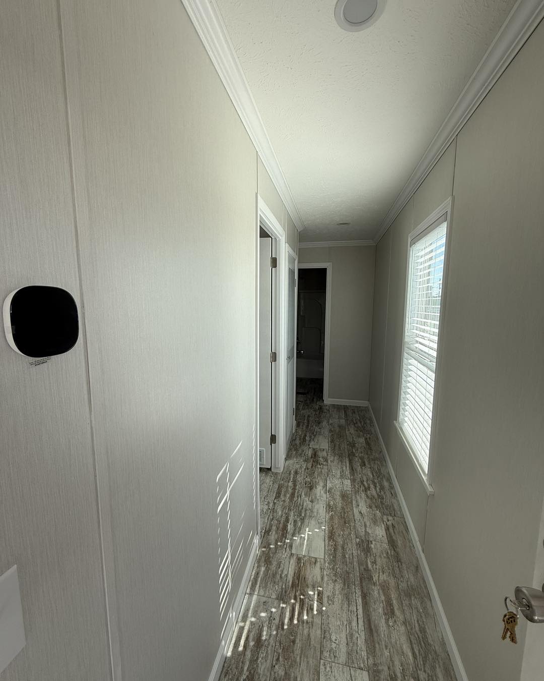 Narrow hallway with light wood flooring, beige walls, a window on the right allowing sunlight in, and a white door at the end. Calm, minimalist ambiance.