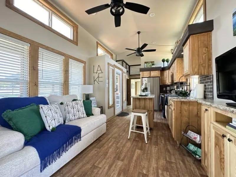 Compact, cozy living space with wooden floors and cabinets. A white sofa with green and blue cushions rests against a wall of windows. Ceiling fans and natural light enhance the airy feel. Kitchen with modern appliances and "LAKE" decor sign add charm.