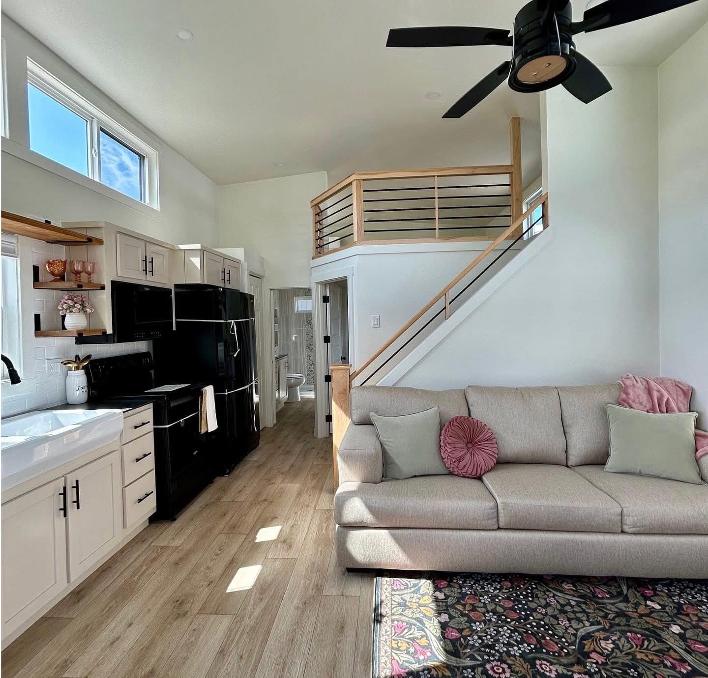 Modern tiny house interior featuring a cozy beige couch with colorful cushions, a black ceiling fan, bright kitchen, loft, and wooden floors.
