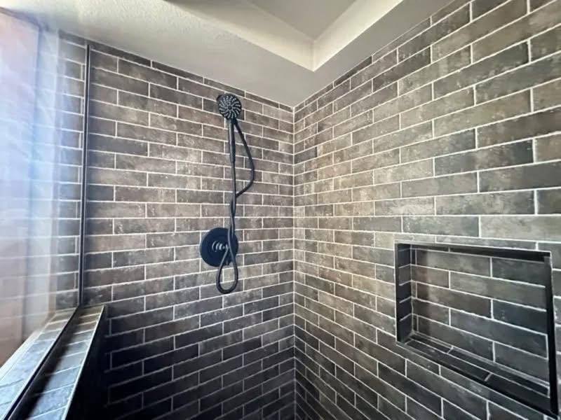Modern shower with dark brown brick tiles, featuring a sleek black showerhead and handle. A recessed shelf is on the right wall. Bright and clean.