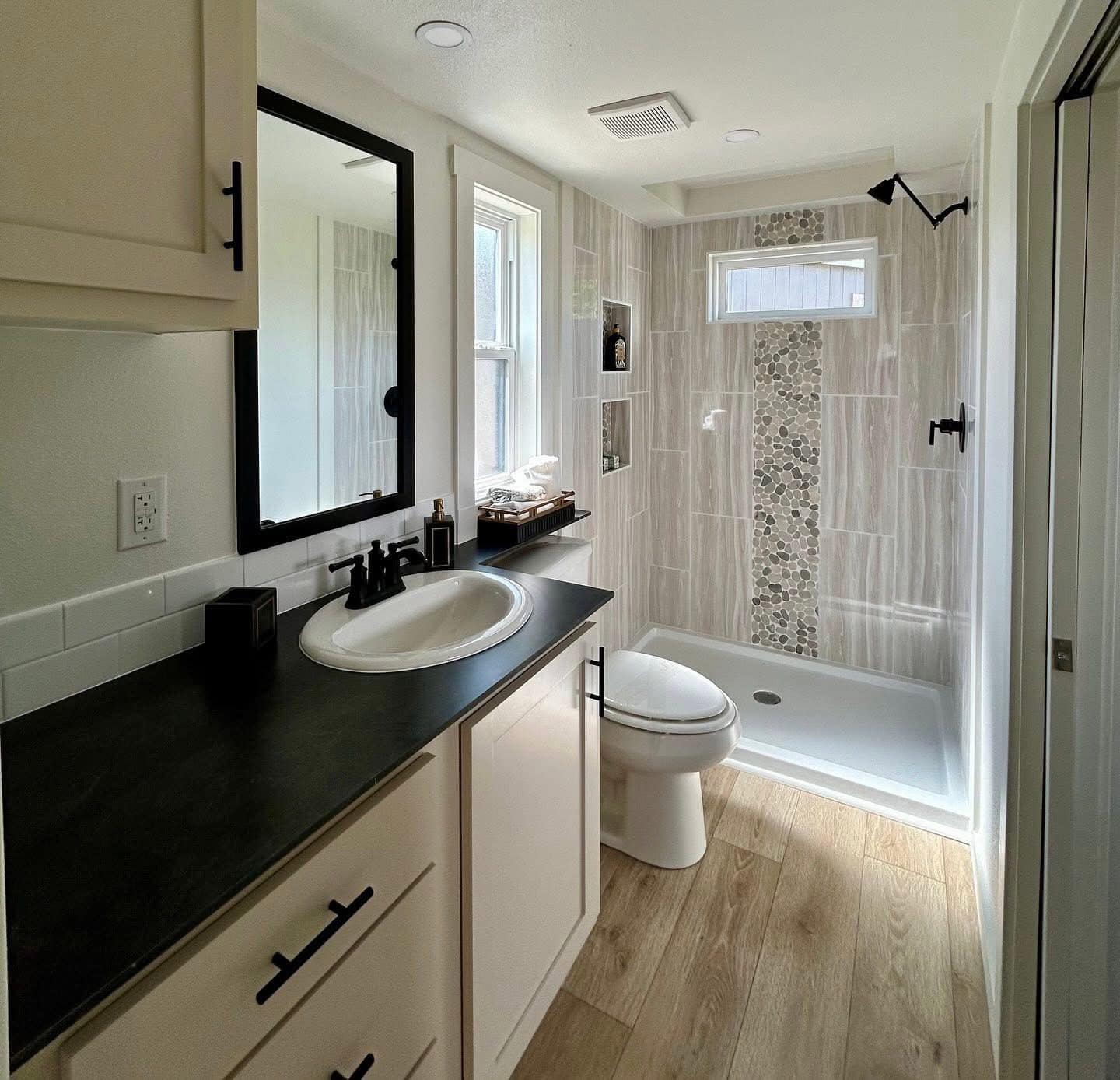 Modern bathroom featuring a sleek black countertop, white sink, and light wood flooring. A shower area with pebble accent tiles exudes a calming vibe.
