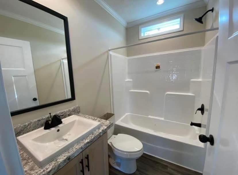 Compact bathroom with white tub-shower combo, beige walls, and wooden floor. Granite-topped sink with black faucet and large black-framed mirror.