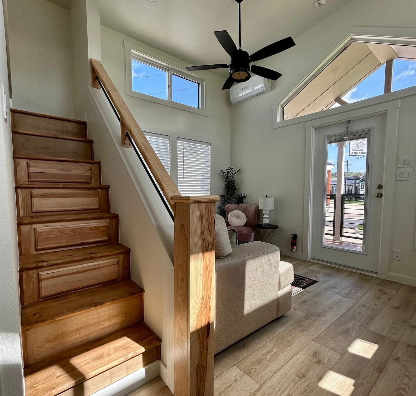 Compact, modern living room with wooden stairs, light wood flooring, and a beige sofa. Large windows and a glass door let in sunlight. Ceiling fan above.