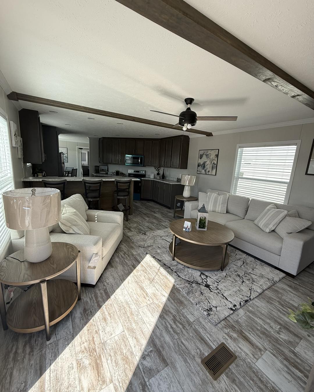 A modern living room with beige sofas, a round coffee table, and wood side tables. Kitchen in the background with dark cabinets and light streaming in. Cozy and inviting ambiance.